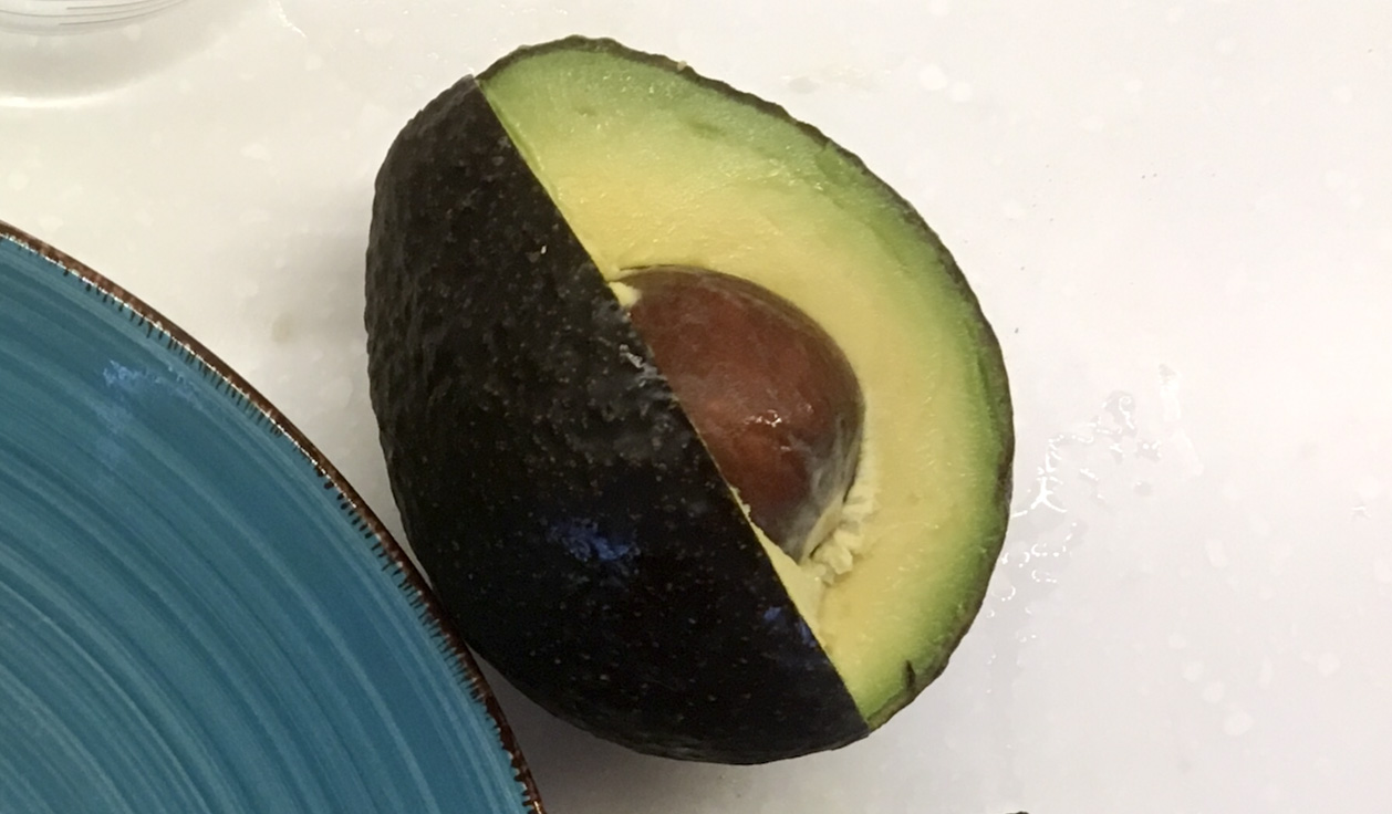 An avoocado with a slice missing.The pit, the white countertop behind,  and the corner of a turquoise plate are visible.
